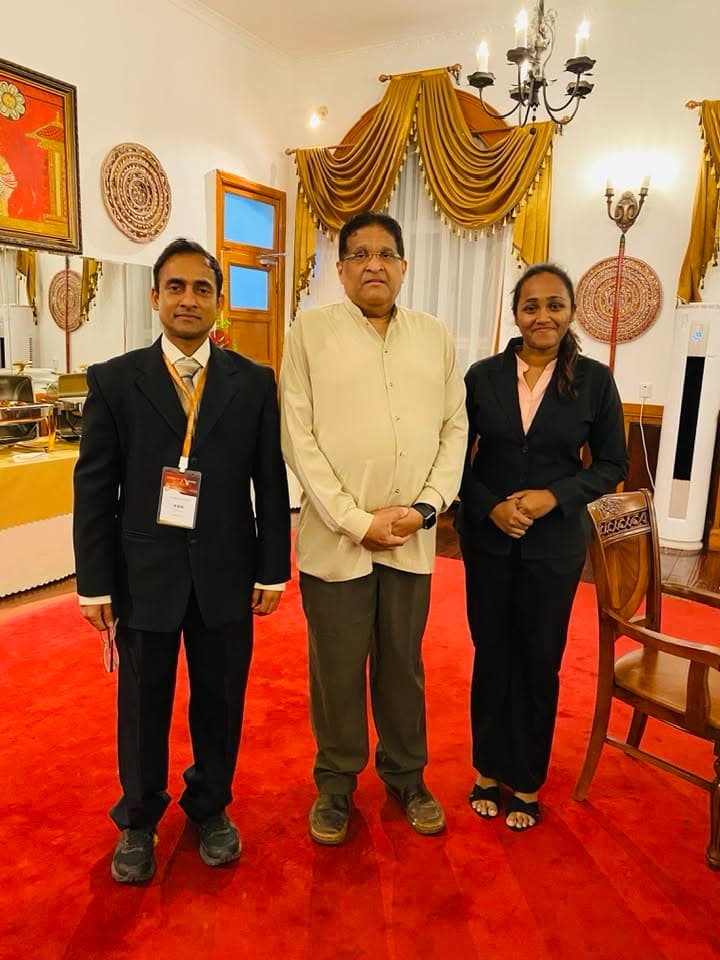Three men stand together on a red carpet in a formal reception room.